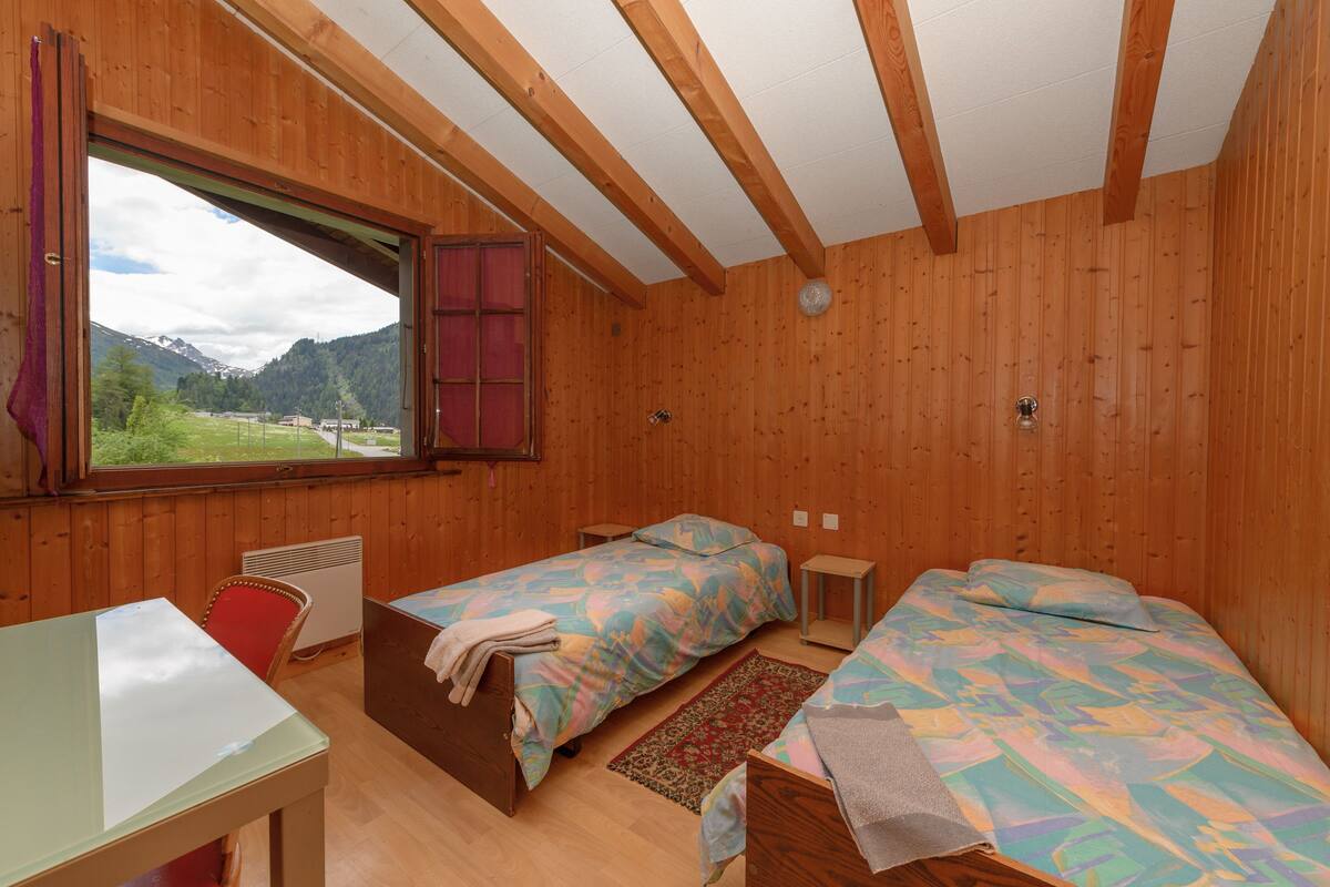 Una habitación con paneles de madera, dos camas, una mesa y un ventanal con vistas a las montañas.