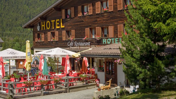 Ein Holzgebäude mit einem Schild, auf dem "HOTEL" und "Cafe Restaurant" auf der Erdgeschossfassade stehen. Vor dem Gebäude befinden sich Terrassentische und -stühle sowie ein Holzzaun.