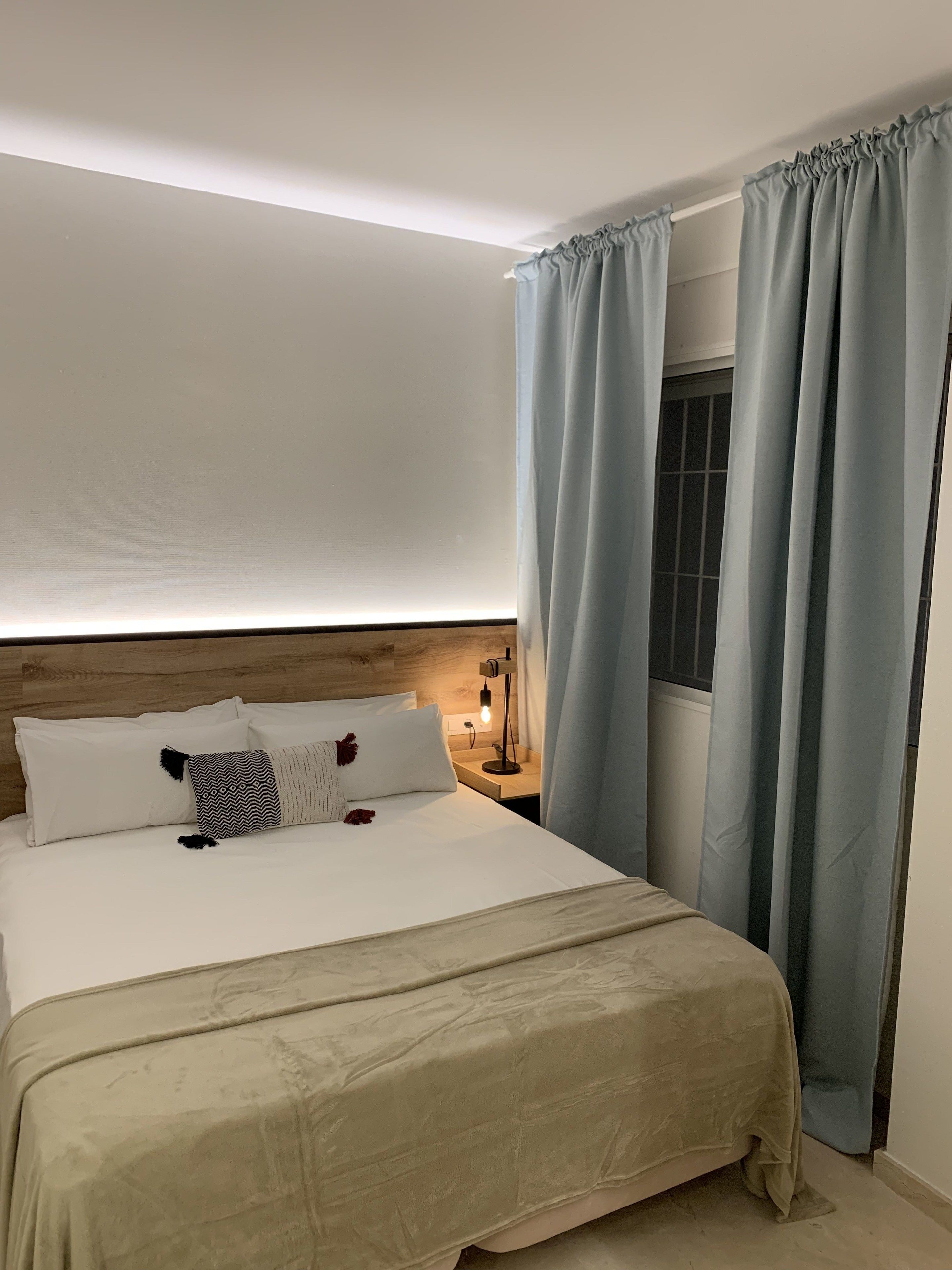 A neatly made bed with a beige bedspread, white pillows, and a patterned throw. There is a wooden headboard, a bedside table with a lamp, and light blue curtains covering a window.