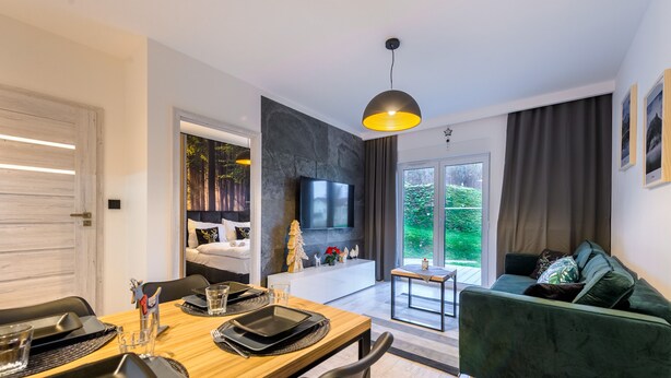 A modern living room with a wooden dining table, a green sofa, a TV mounted on a stone wall, and a view of the outdoors through large windows. - Izerska Przygoda - Apartamenty 5d (Swieradow-Zdroj)