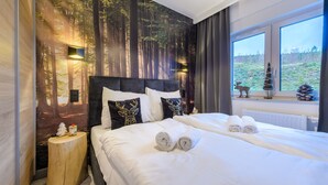A bedroom with a forest-themed wallpaper, a large bed with white linens, a nightstand with a lamp, and a window with a view of a field and a reindeer statue. - Izerska Przygoda - Apartamenty 5d (Swieradow-Zdroj)
