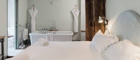 A hotel room with a large bed, a wooden headboard, a white bathtub, and two white robes hanging on the wall.