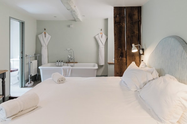 A hotel room with a large bed, a wooden headboard, a white bathtub, and two white robes hanging on the wall.