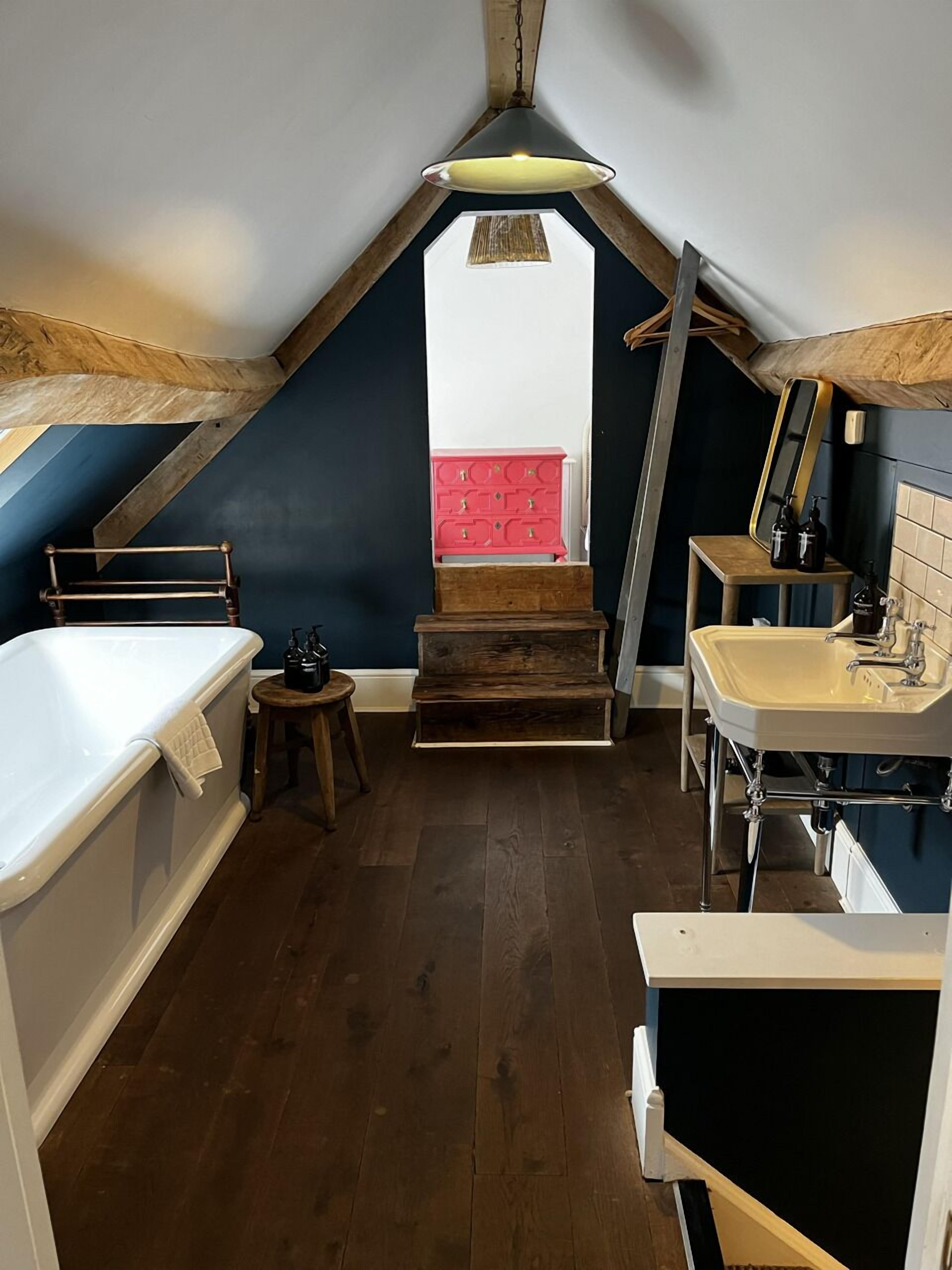 Un dormitorio acogedor en el ático, con techo inclinado, una bañera blanca, un banco de madera, un aparador rojo y un lavabo blanco.
