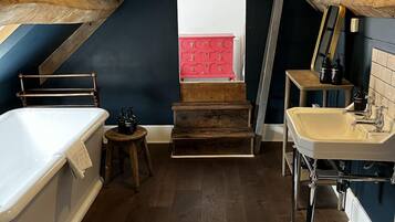 Un dormitorio acogedor en el ático, con techo inclinado, una bañera blanca, un banco de madera, un aparador rojo y un lavabo blanco.
