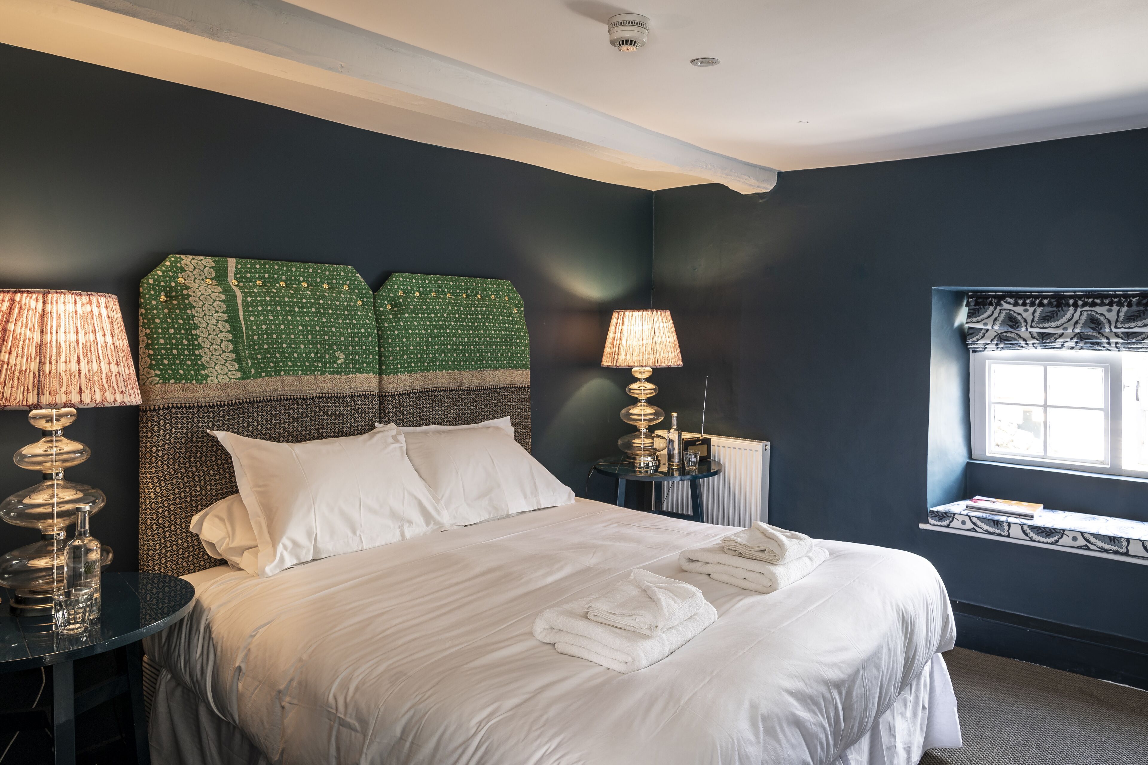 A neatly made bed with white linens and towels, a headboard with a green patterned design, two bedside lamps, and a window with a view.