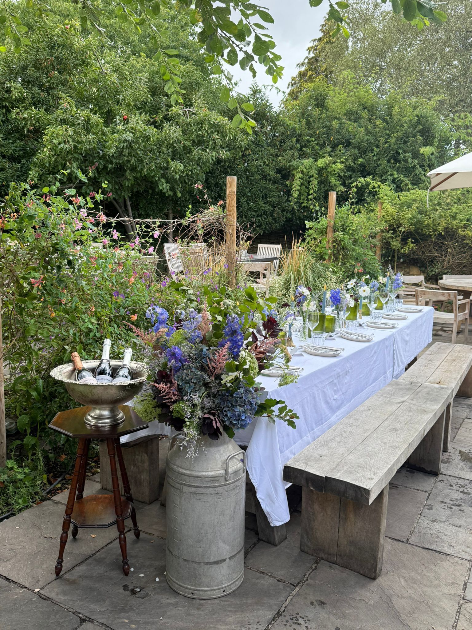 An outdoor dining area with a long table set for a meal, surrounded by lush greenery and a variety of flowers.
