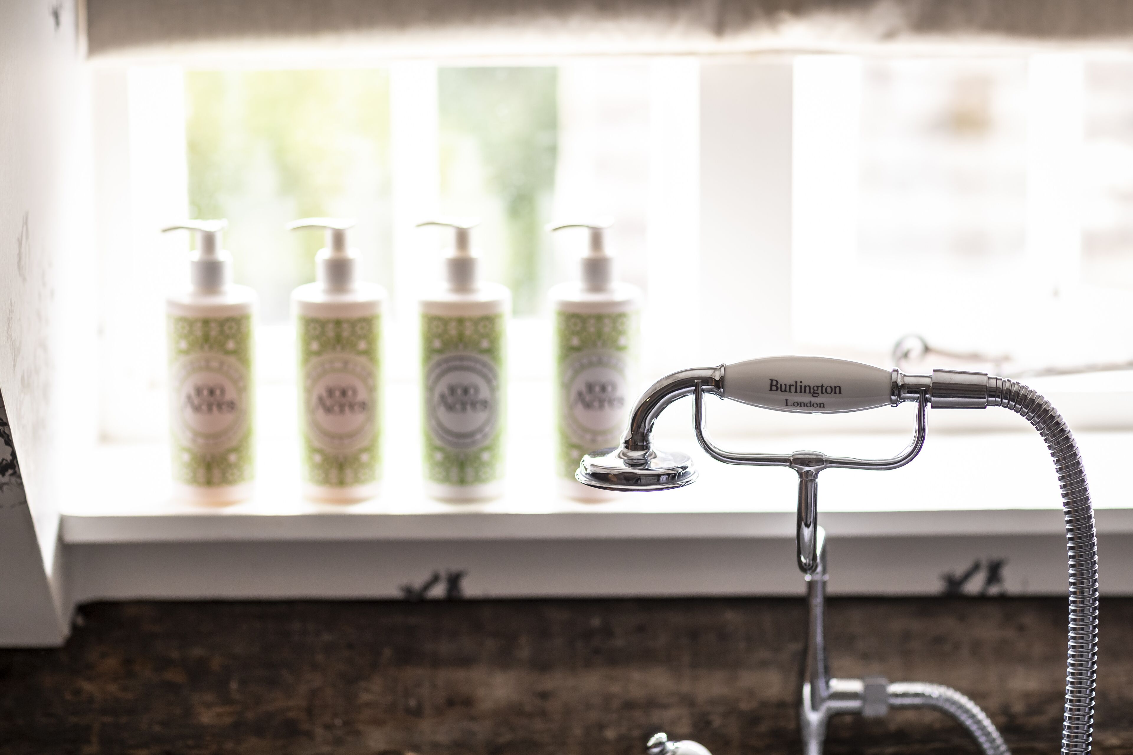 Een badkamer met een douchekop met de tekst “Burlington London” en vier flessen “100% Natuurlijk” shampoo op een plank.