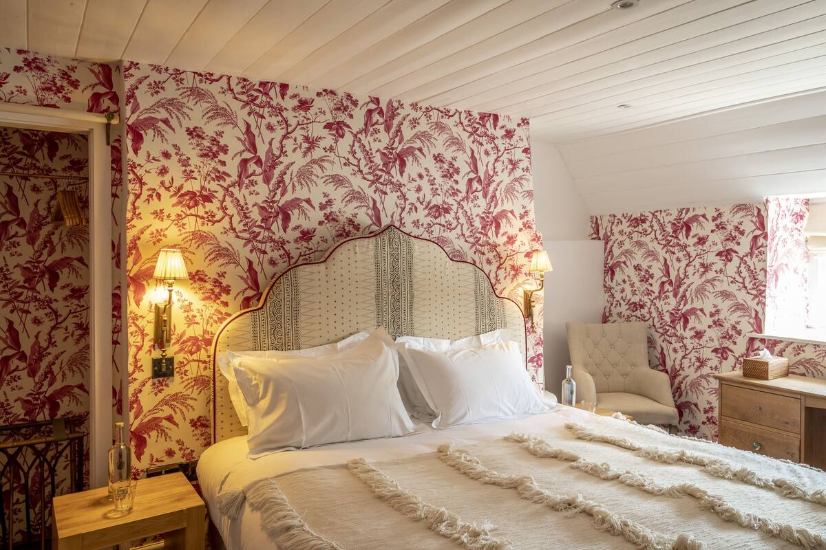 A bedroom with floral wallpaper, a bed with white linens, a nightstand with a bottle, and a chair.