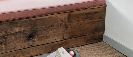 Habitación de esquina con un banco de madera, una ventana con persiana estampada y una cama para mascotas con objetos encima.