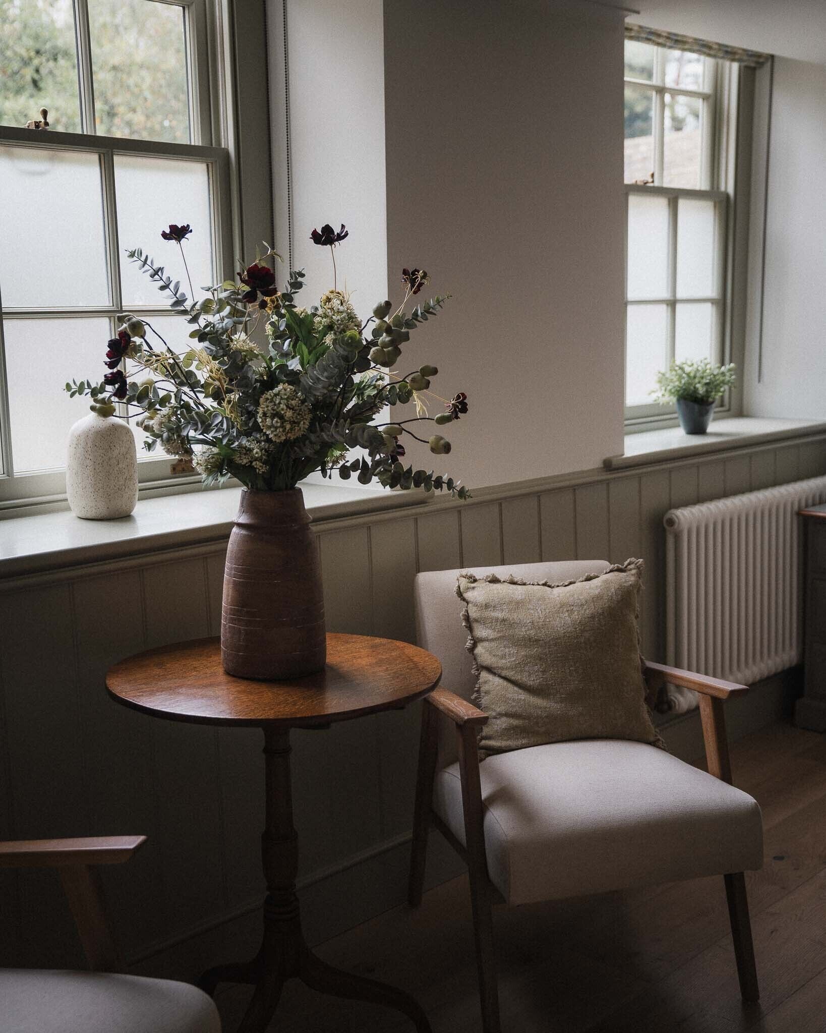 A room with a round wooden table, a chair, a vase with flowers, and a window with a view of trees.
