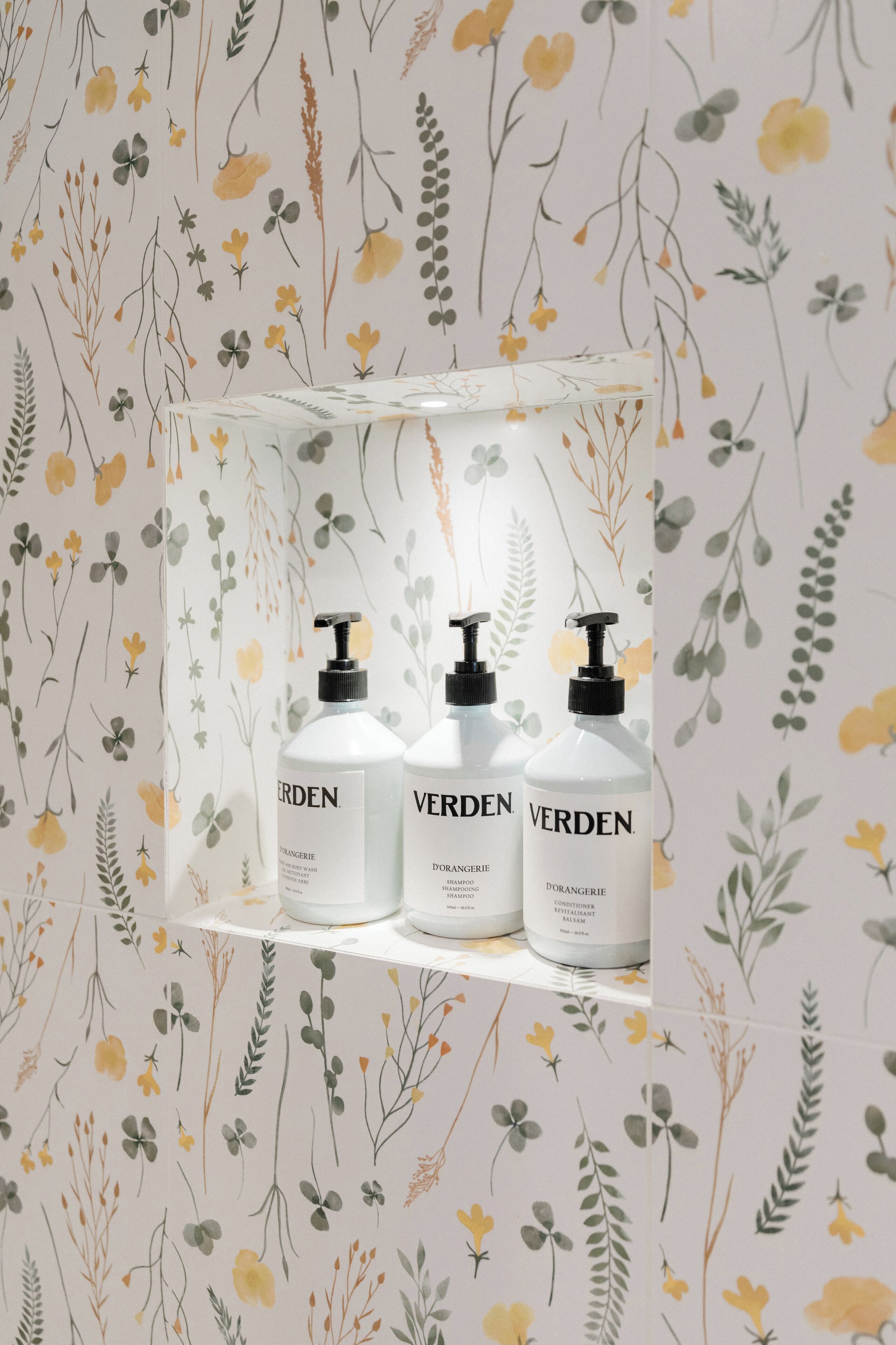 Three bottles of Verden body wash on a shelf against a floral wallpaper background.