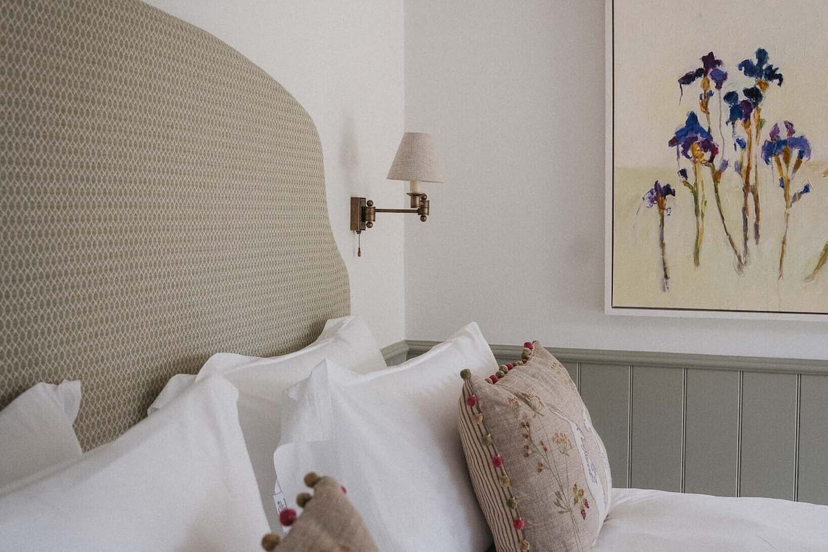 A neatly made bed with white linens and decorative pillows, a wall-mounted lamp, and a painting of flowers.