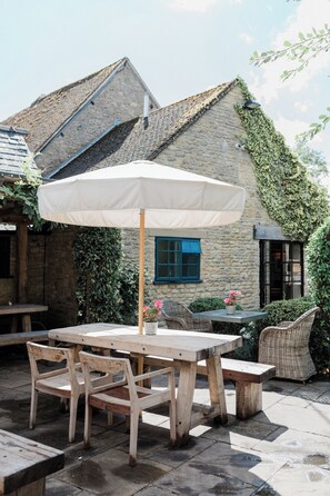 An outdoor patio with a wooden table and chairs, a white umbrella, and a stone building with ivy. - The Swan Inn (Chipping Norton)
