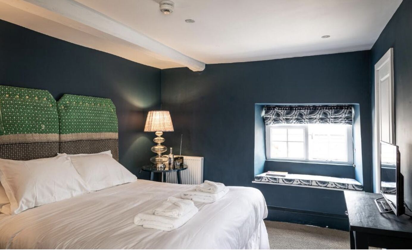 A hotel room with a large bed, a bedside table with a lamp, a window with a patterned valance, and a view of the outside.
