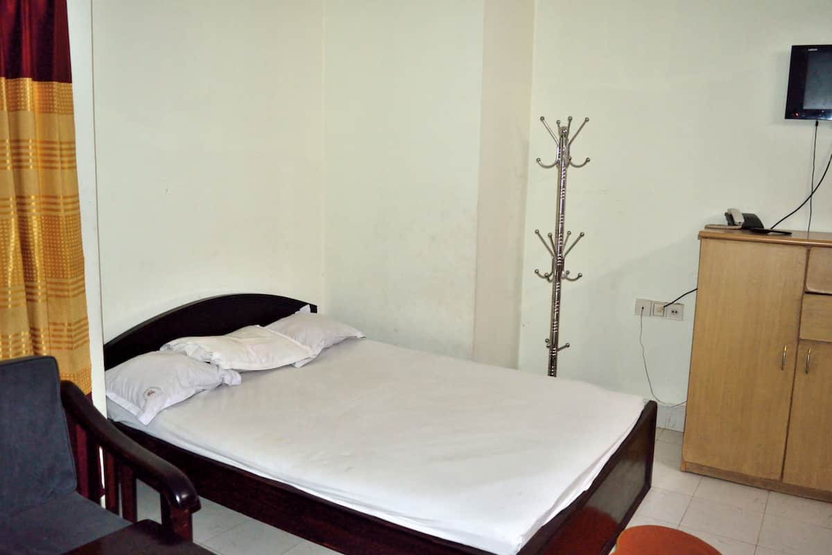 A single bed with white linens, a wooden headboard, a coat rack, a television mounted on the wall, and a wooden wardrobe.