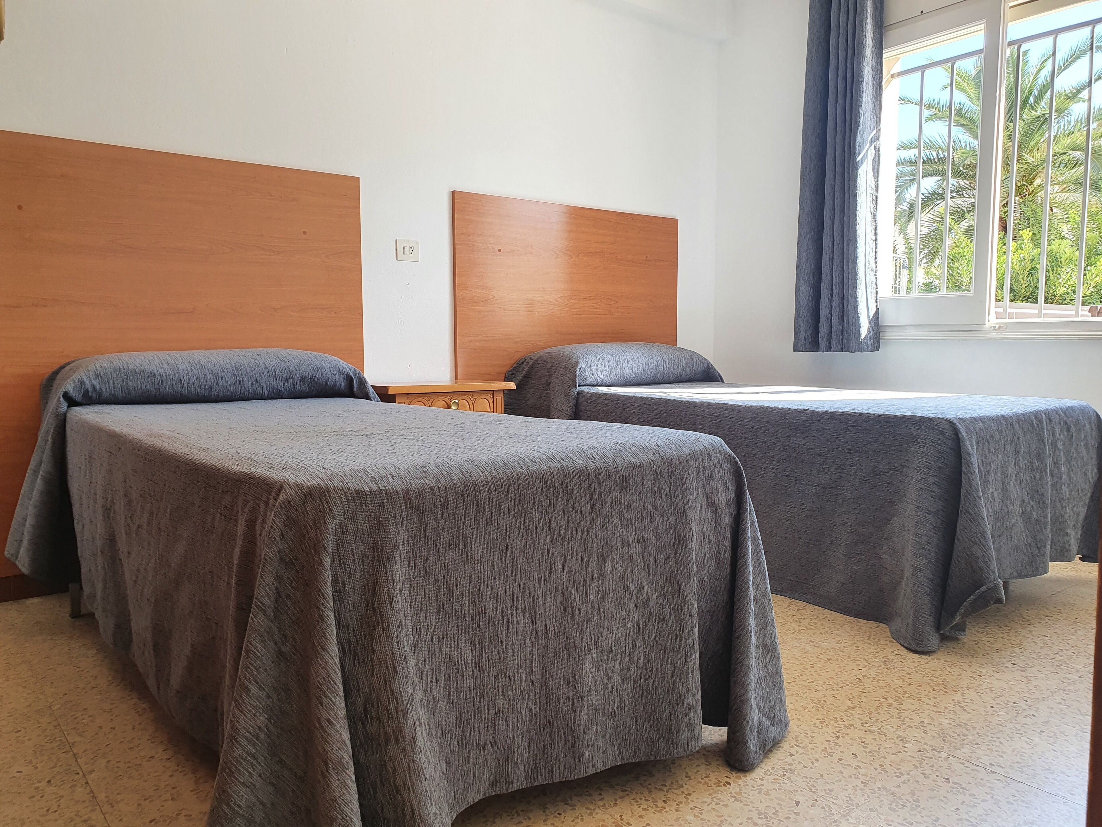 Une chambre d’hôtel avec deux lits simples, une tête de lit en bois, une petite table de chevet en bois et une fenêtre donnant sur des palmiers.