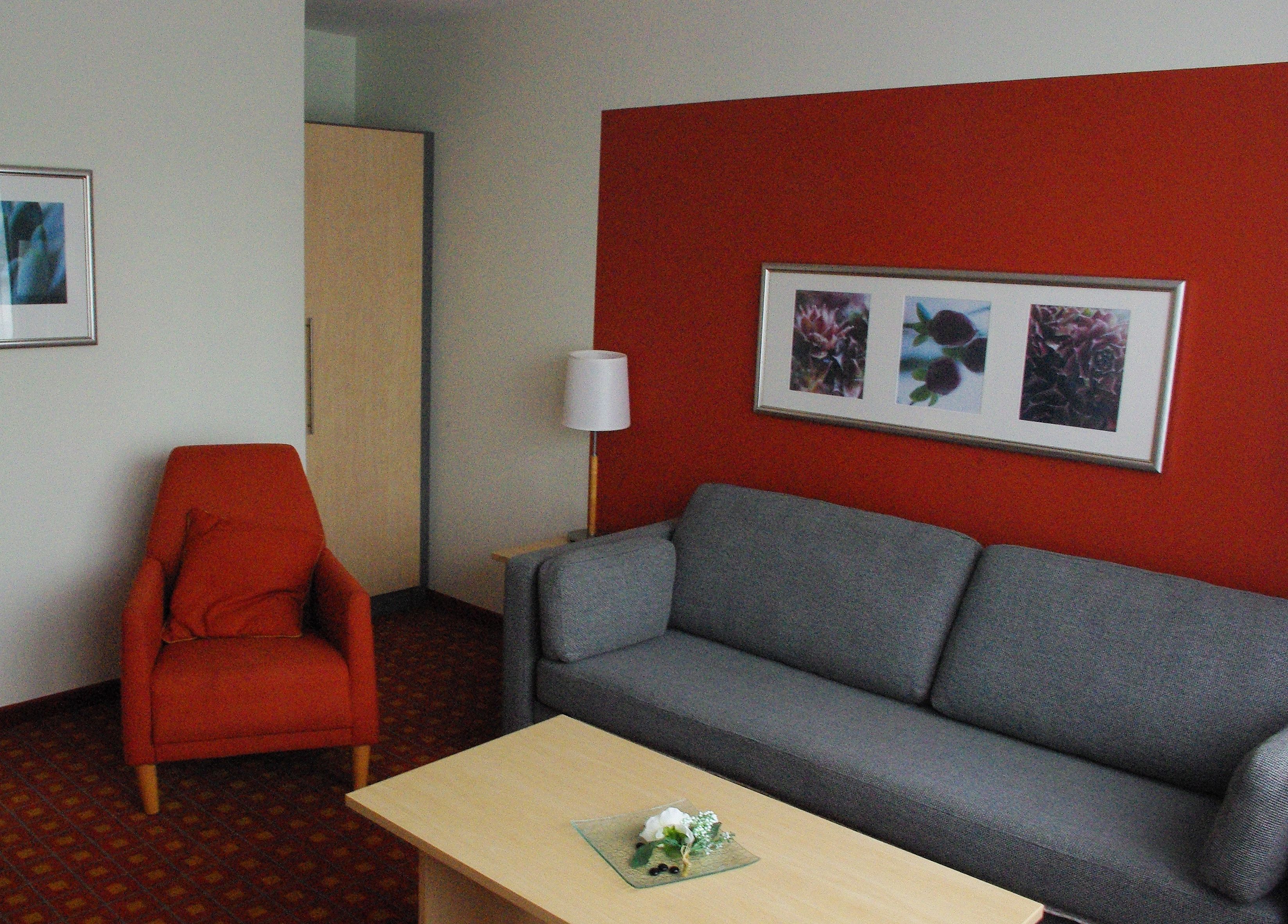 Ein Hotelzimmer mit roter Akzentwand, einem grauen Sofa, einem roten Sessel, einem kleinen Tisch mit einer Blume und einem gerahmten Bild über dem Sofa.