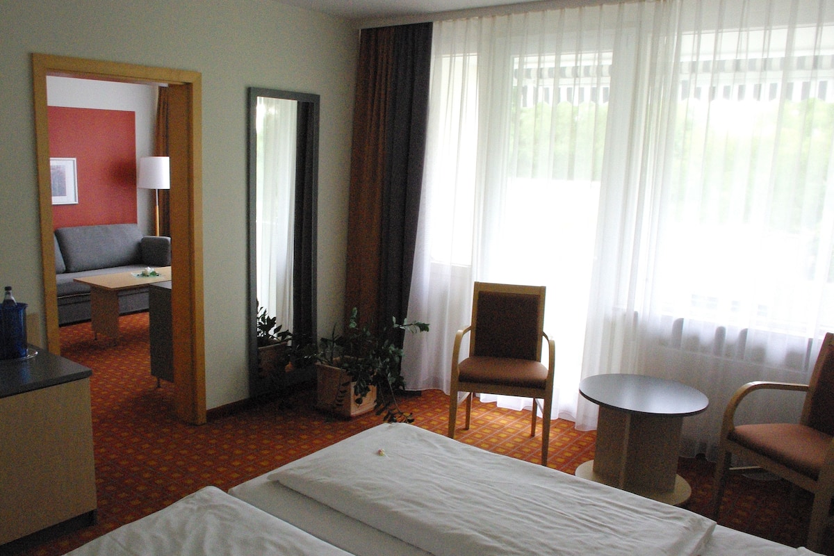 A hotel room with a bed, a desk, a chair, a small table, and a potted plant.