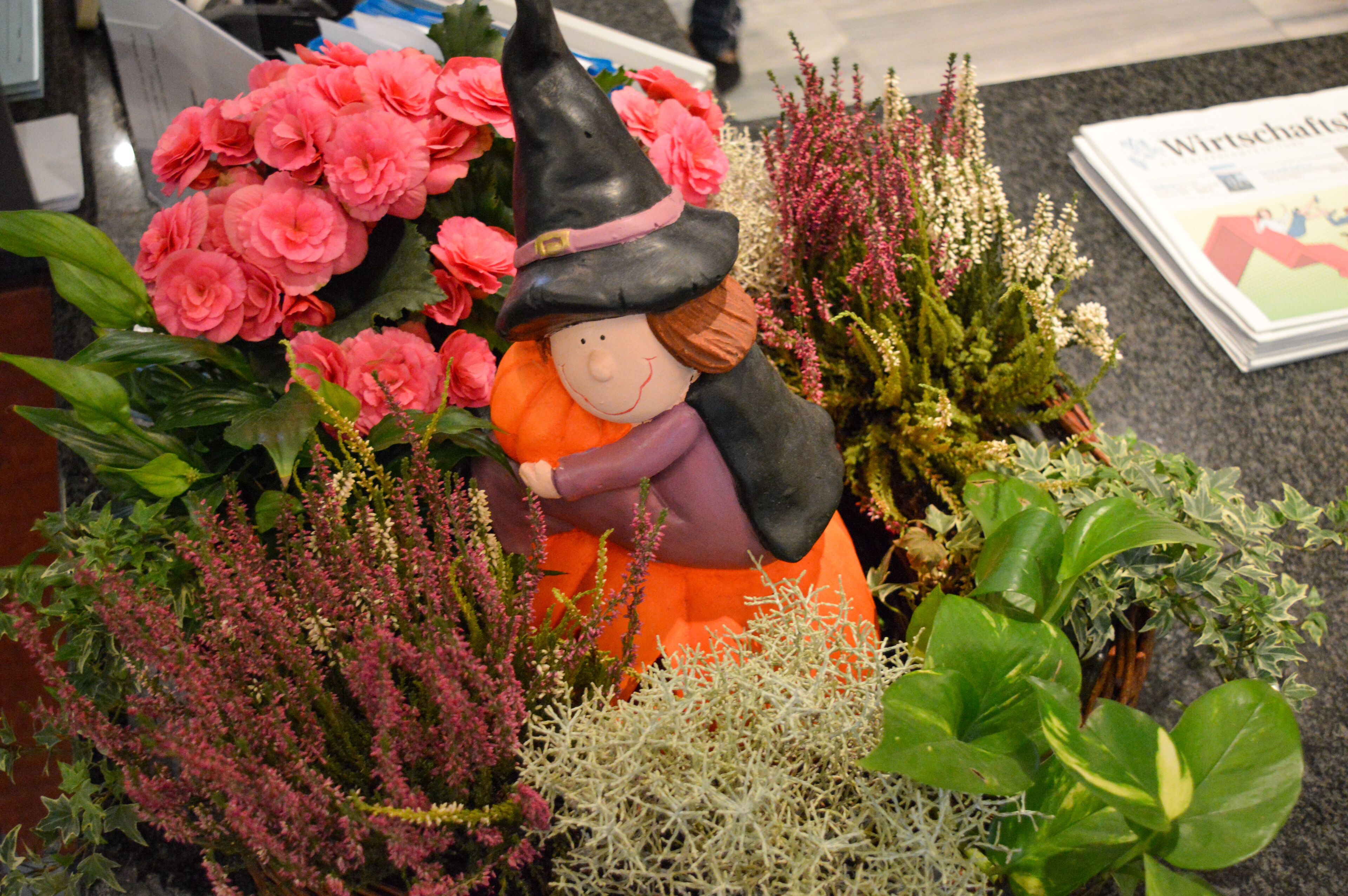 Eine dekorative Hexenfigurine umgeben von verschiedenen Pflanzen und Blumen.