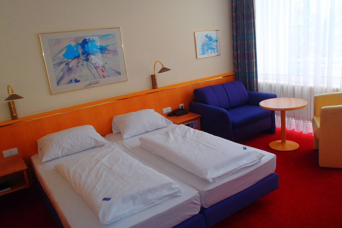 A hotel room with two beds, a blue armchair, a small round table, and two paintings on the wall.