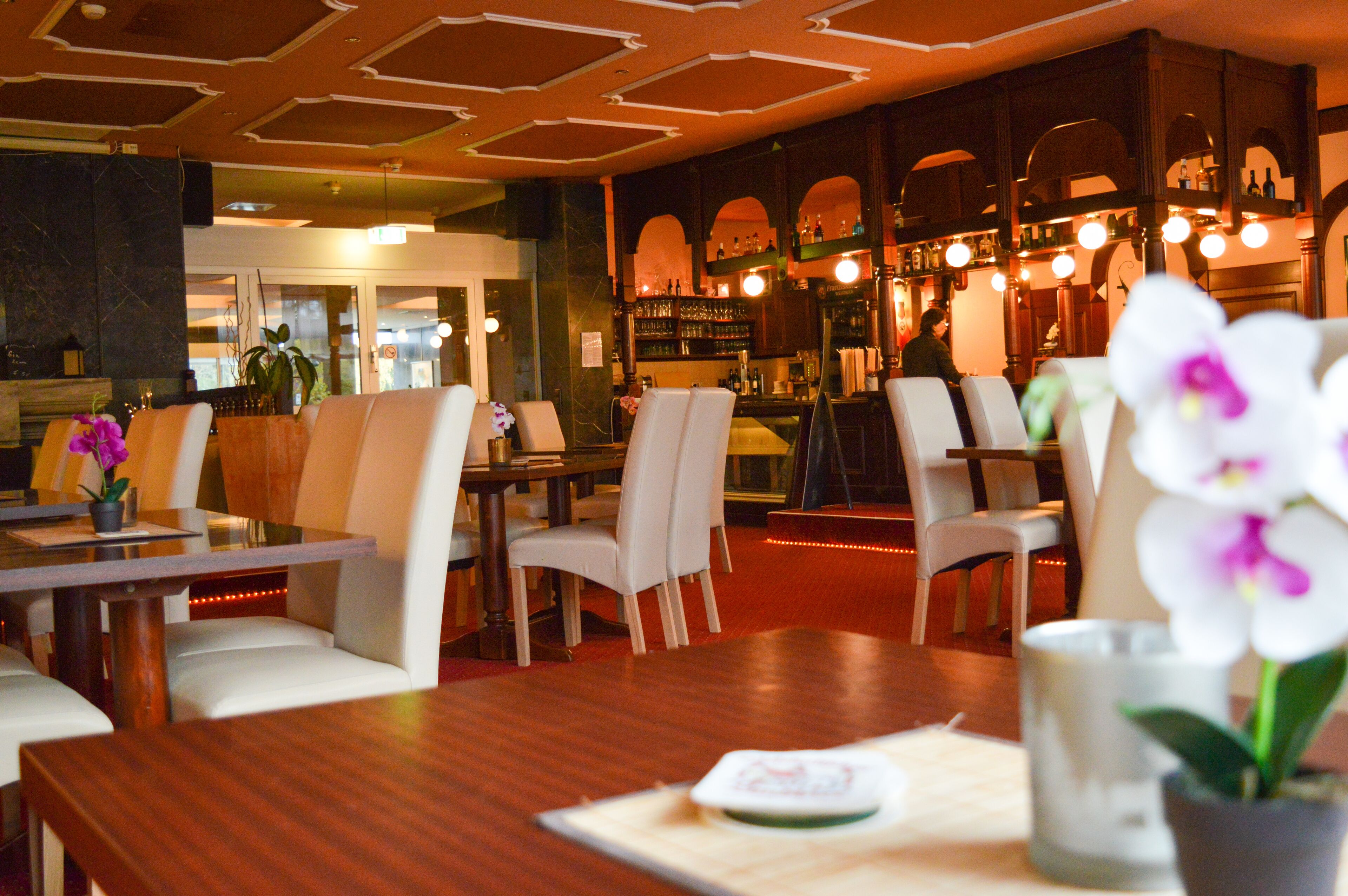Ein Restaurant mit Holztischen und weißen Stühlen, einem Barbereich mit Flaschenregalen und einer dekorativen Decke.