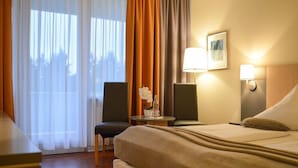 A hotel room with a bed, two chairs, a small table with a lamp, and a window with curtains.