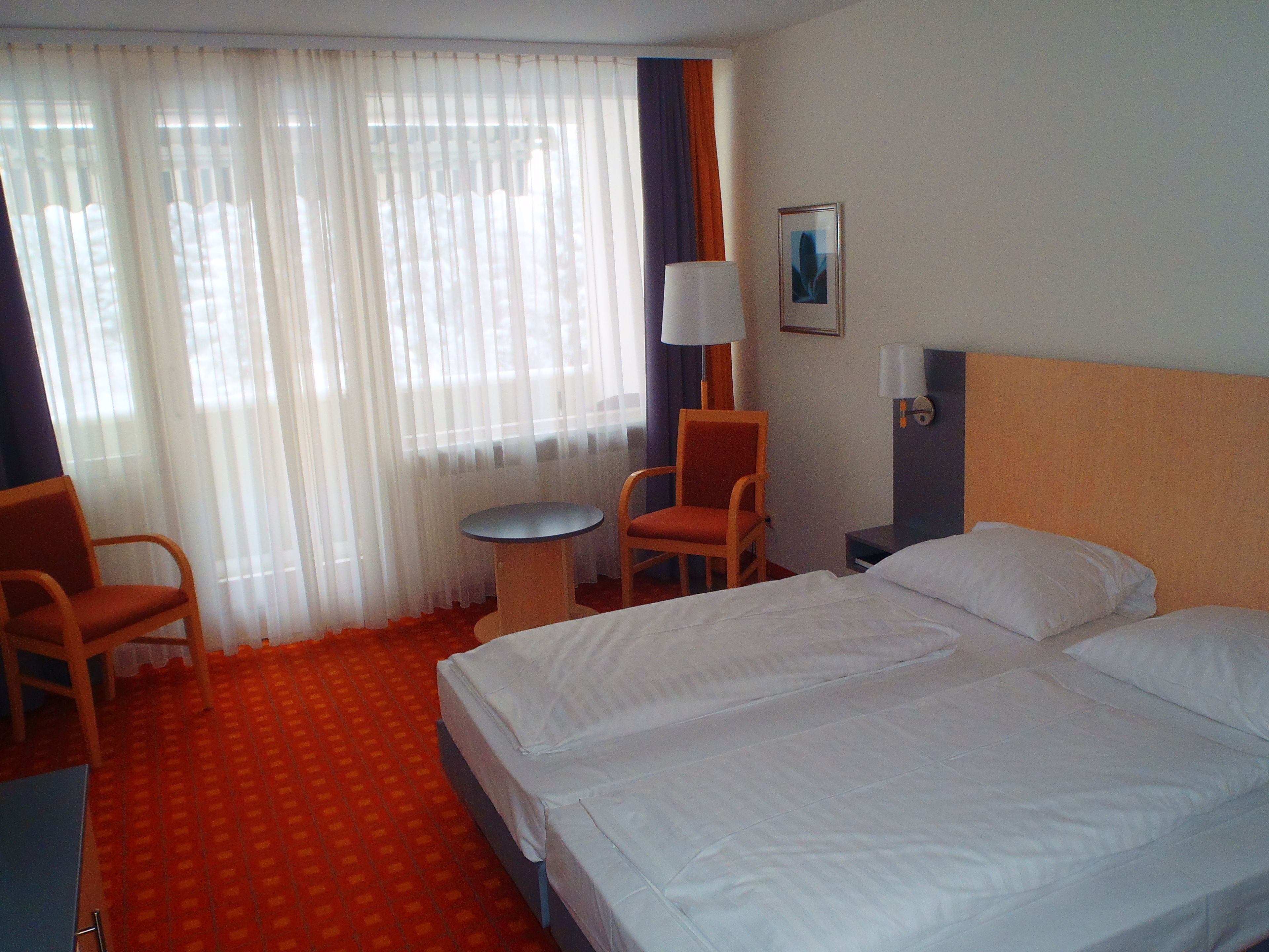 Camera d'albergo con un letto, due sedie, un tavolino, una lampada e una finestra con tende.