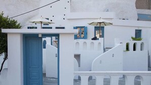 A white building with a blue door, white walls, and blue shutters. There are umbrellas and a small plant on the roof. - Stoa Cave Villas (Santorini)