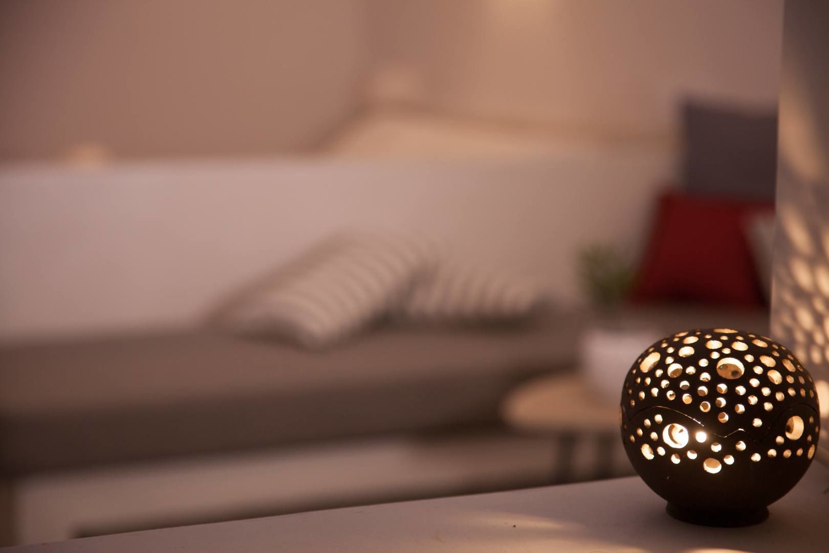A spherical lamp with a perforated design on a table in a room with a bed and pillows in the background.
