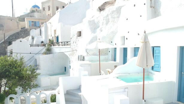 A white-walled building with a pool, blue shutters, and a staircase leading to a higher level. - Stoa Cave Villas (Santorini)