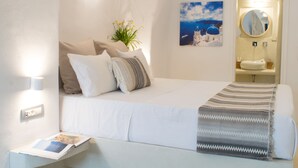 A neatly arranged hotel room with a bed, a bedside table with a magazine, a wall-mounted mirror, a sink, and a painting of a blue-domed building. - Stoa Cave Villas (Santorini)