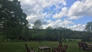 An outdoor seating area with wooden tables and chairs, surrounded by trees and a grassy field.