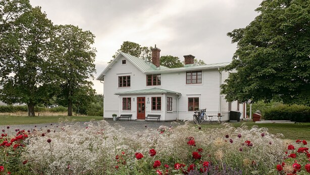 A white two-story house with a green roof, surrounded by a garden with red and white flowers.