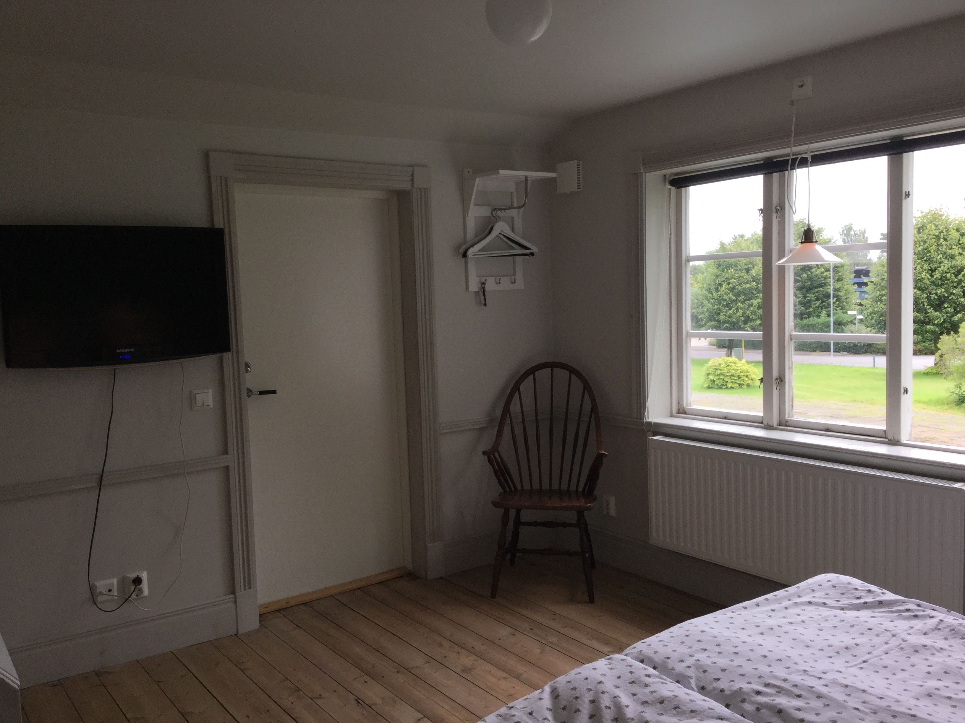 A room with a bed, a chair, a TV mounted on the wall, and a window with a view of the outdoors.