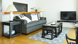 A modern living room with a grey sofa, two side tables with lamps, a coffee table, a flat-screen TV, and a patterned rug. - Winbell Okinawa Nago Coral View (Nago)