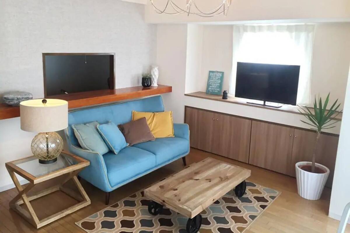 Sala de estar com sofá azul, mesa de centro de madeira, mesa lateral de vidro com luminária, televisão de tela plana e planta em vaso.