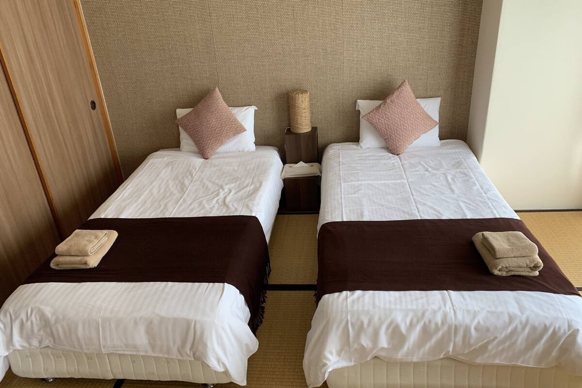 Duas camas de solteiro com roupa de cama branca e cabeceiras marrons, uma cabeceira trançada e uma pequena mesa de cabeceira com abajur.