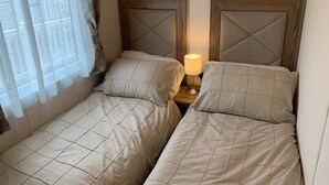 Habitación doble con dos camas, una mesita de noche con lámpara, cortinas translúcidas y vistas a edificios.