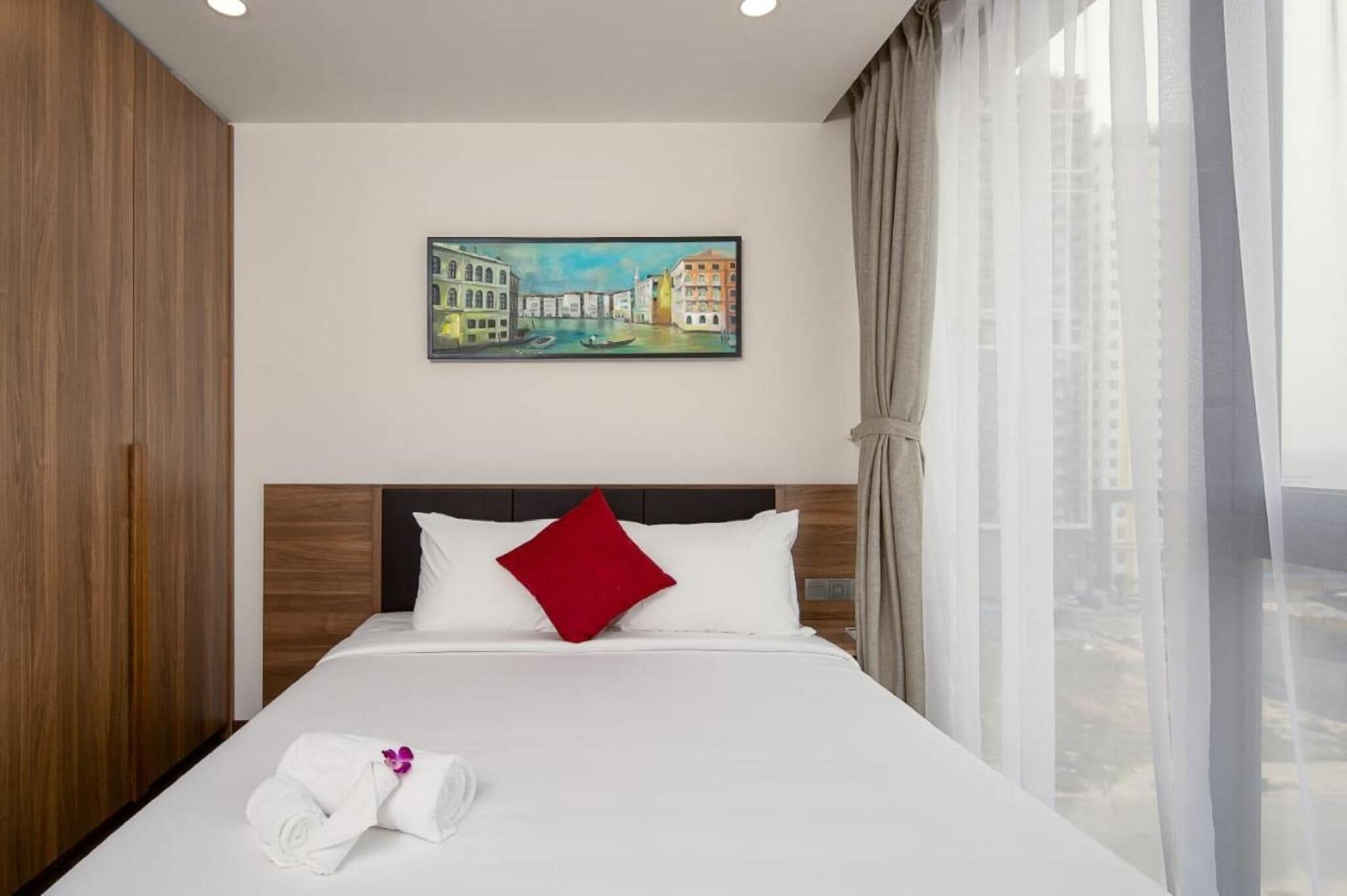A hotel room with a bed, a painting of a cityscape, a red pillow, and a window with curtains.