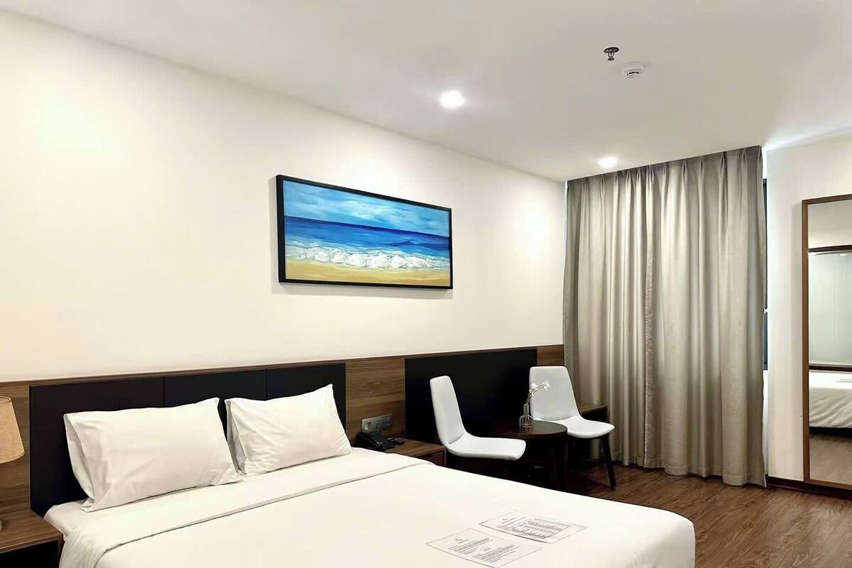 Une chambre d’hôtel avec un grand lit, un bureau, une chaise, un tableau représentant une plage et une fenêtre avec des rideaux.