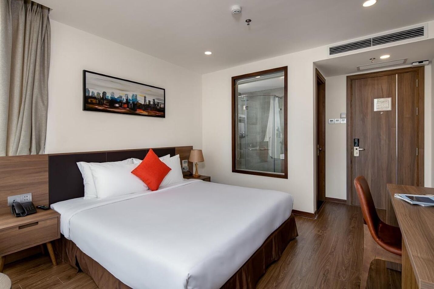 A hotel room with a bed, a desk, a chair, a mirror, a painting, and a door.