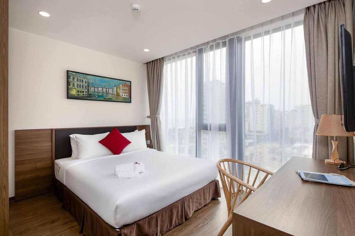 Une chambre d’hôtel avec un grand lit, un bureau, une chaise, un tableau au mur et une vue sur la ville par la fenêtre.