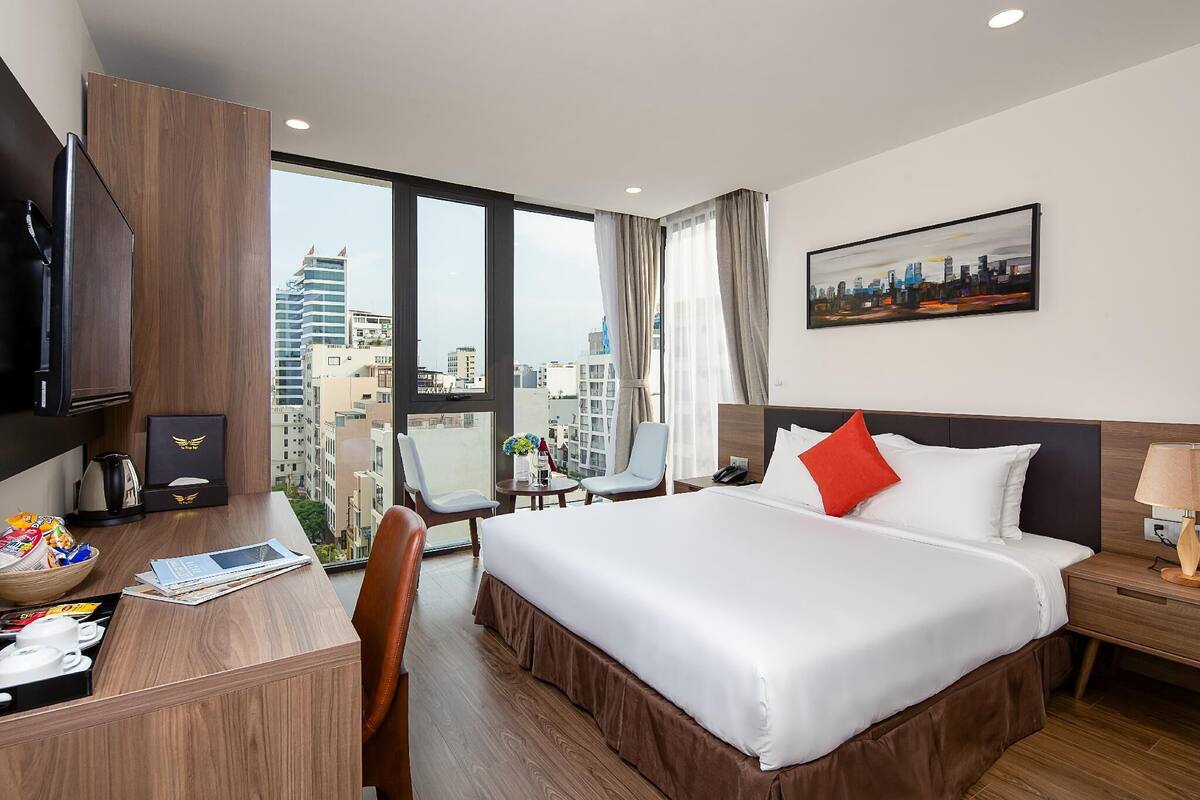 Une chambre d’hôtel avec un grand lit, un bureau avec un ordinateur, une chaise, une petite table avec un vase et une vue sur la ville à travers de grandes fenêtres.