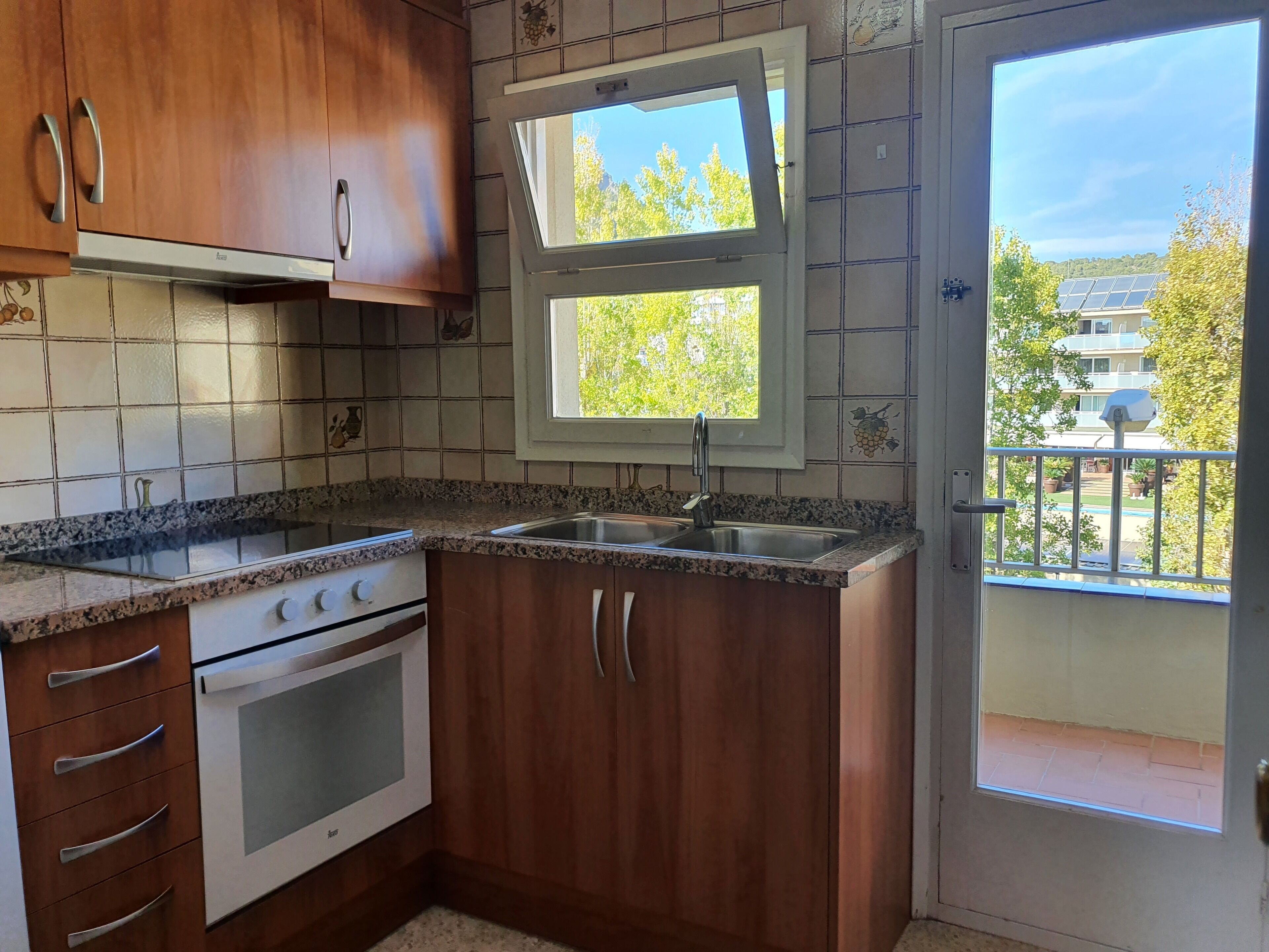 Una cocina compacta con armarios de madera, un lavamanos de acero inoxidable y una ventana con vistas a un balcón con paneles solares.