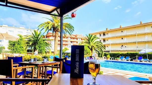 Un bar junto a la piscina con una cerveza, asientos al aire libre y vistas a una piscina y edificios de hotel.