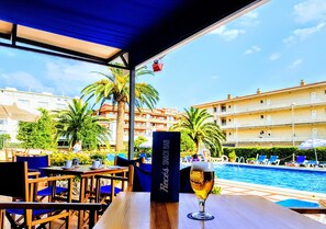 A poolside bar with a glass of beer, outdoor seating, and a view of a swimming pool and hotel buildings. - Payet Apartments 2-4 Personas (Torroella de Montgrí)