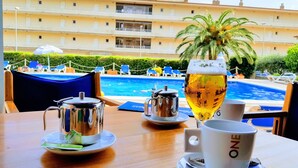 A table with a teapot, a cup labeled "ONE", and a glass of beer, overlooking a pool with lounge chairs and a building in the background.