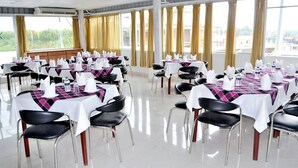 Ein Konferenzraum mit mehreren Tischen und Stühlen, großen Fenstern mit Vorhängen und Blick auf Grünflächen.