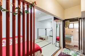 Una habitación tradicional japonesa con suelo de tatami, una cama baja de madera con ropa de cama roja, una pequeña mesa y una puerta corredera que da a un baño con lavabo y bañera.